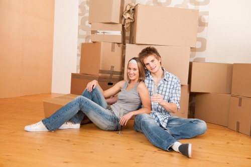 A young couple sitting on a polished wooden floor in a room with a plain white wall, surrounded by multiple large cardboard boxes, some sealed with brown packing tape and others partially open, suggesting a move or packing process. The woman, wearing a grey tank top and light blue jeans, is reclining against the man with her legs stretched out and a broad smile, while the man, dressed in a blue and white checkered shirt and blue jeans, sits comfortably with his arm around her shoulder. The background features a neat stack of boxes of varying sizes, positioned against the wall, indicating an ongoing house relocation. Natural lighting illuminates the scene, creating a warm and inviting atmosphere typical of house removals in the Chiswick area, as part of professional services offered by Chiswick Man with Van. The room has a minimalistic look, with no additional furniture visible, emphasizing the removal staging with a focus on boxes and packing items, representative of a house moving situation in a suburban London setting.