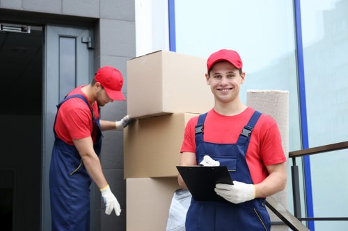 Two professional removals workers from Chiswick Man with Van are engaged in a house relocation outside a modern residential building, which features large glass windows and a grey exterior wall. The worker on the left, dressed in a red cap, red t-shirt, blue overalls, and white gloves, is lifting or adjusting a large cardboard box stacked on top of two other boxes near the building's entrance. The second worker, in similar uniform with a red cap, red t-shirt, and blue overalls, stands in the foreground holding a black clipboard and smiling at the camera, suggesting a positive and organized moving process. Behind them, the boxes appear to be sturdy, made of brown cardboard, and are used for packing household items. The setting indicates a residential street or driveway in the Chiswick area, with natural daylight illuminating the scene, emphasizing the professional approach taken by the team to facilitate house removals and sustainable recycling practices. The focus remains on the clear, detailed depiction of the workers, their equipment, and the environment to support local moving and recycling services.