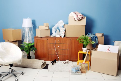 A living room scene with a blue wall background shows various household items and moving supplies. In the centre, a wooden sideboard is topped with several cardboard boxes, some open and others sealed, along with a small potted plant, a plastic bag, and a cloth draped over a box. To the left, a large white plastic bag or wrapping material partially covers an item, while a rolled-up rug with a beige fabric finish lies on the floor nearby. A black handheld vacuum or small appliance rests in front of the sideboard, connected to a cord. To the right, a yellow step stool and a small orange wicker basket are positioned beside a large cardboard box, with some loose items and smaller boxes scattered around. There are two potted plants, one on each side of the sideboard, adding greenery to the scene. The floor is tiled, and natural light illuminates the space, suggesting a daytime setting in a residential room during a house move or packing process, consistent with house removal services in the Chiswick area.