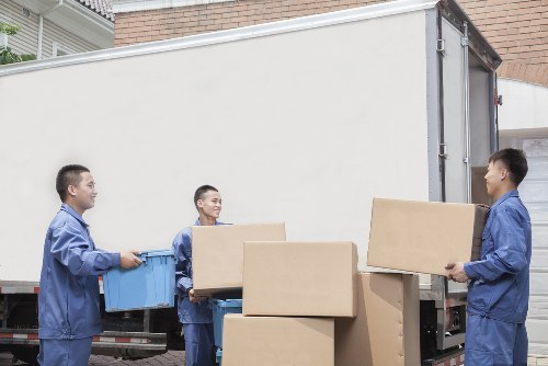 Three men dressed in blue work uniforms are engaged in loading cardboard boxes and plastic crates onto a large white removals truck parked on a residential street, possibly in the Chiswick area. One man on the left is holding a blue crate, while the middle man is carrying a cardboard box and the rightmost man is handing another box to him. The boxes vary in size, some appearing to be filled with household belongings, and are made of brown cardboard with visible creases and tape. The truck's rear doors are open, revealing the interior space for loading, and the backdrop includes a brick wall and a residential building, suggesting a typical London suburb environment. The scene is well-lit with natural daylight, emphasizing the professional activity associated with house removals by Chiswick Man with Van, part of the removals service category, in the area's local context. The focus is on the organized effort of the team as they prepare items safely for transportation, aligning with health and safety policies relevant to moving house in the W4 postcode area.