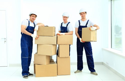 Three professional movers from Chiswick Man with Van, dressed in navy blue uniforms and white helmets, stand inside a bright, modern room with white walls and large windows. They are positioned around a stack of medium-sized cardboard boxes, carefully holding additional boxes to load or unload. The boxes are sealed with packing tape and are of varying sizes, some stacked directly on the floor, others held by the movers. The flooring appears to be light-colored laminate or vinyl, contributing to the clean, minimalistic environment, which is typical of a residential property in Chiswick or nearby London postcode areas. Natural daylight streams through the windows, illuminating the scene and creating a professional and reassuring atmosphere for house removals. The movers are focused, prepared, and demonstrate careful handling of the household items, emphasizing the company's commitment to secure and efficient relocation services in the local area.