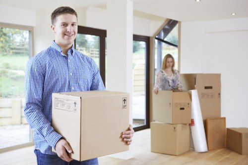 A young man in a blue checkered shirt is standing inside a well-lit residential space, holding a medium-sized cardboard box with shipping labels. Behind him, there is a woman with long blonde hair, wearing a patterned blouse, also with cardboard boxes around her, some stacked on the wooden floor and one leaning against a wall. The interior features large windows allowing natural light to fill the room, revealing a patio or garden area outside. The scene depicts a house removal or moving process, with household items such as boxes and minimal furniture visible, reflecting a professional house removals service in the Chiswick area. The overall environment appears clean and organized, with unpacked items and packing materials present, set within a modern home with white walls and bright natural lighting typical of a residential property during a move.