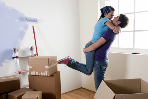 A young couple engaged in a move within a bright, modern room that features white walls and a large window allowing natural light to illuminate the space. The man, wearing a purple T-shirt and blue jeans, is holding the woman, dressed in a blue top and dark jeans, in a lifting embrace. The woman has her arms around the man’s shoulders, with her legs slightly lifted off the ground as they share a joyful moment. In the background, there are several cardboard boxes of various sizes stacked against the wall, some sealed with packing tape, indicating they are in the process of being packed or moved. To the left, a roll of packing paper is partially unrolled, attached to a wall-mounted dispenser, and a red-handled lifting strap is visible, suggesting the use of moving aids. The flooring is light-colored wood, typical of a residential home in Chiswick, London, and the scene captures a moment of packing or preparing for house removals, with a focus on the person's activity of loading or unpacking belongings, consistent with services provided by Chiswick Man with Van in this area.