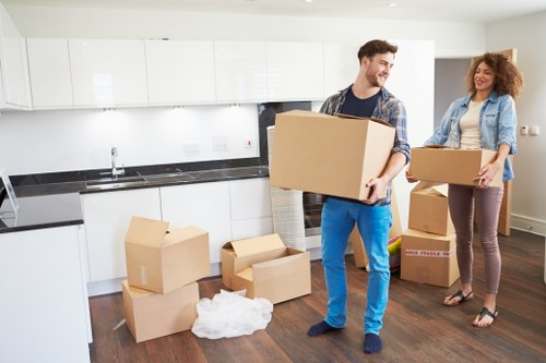 An interior view of a modern kitchen with white cabinets, a black countertop, and a stainless steel sink. Two young adults, a man and a woman, are in the process of unpacking or preparing to move belongings; the man is lifting a cardboard box, wearing a plaid shirt and blue jeans, while the woman, dressed casually in a denim jacket and brown pants, is holding another box and smiling. Several cardboard boxes, some sealed and others open, are scattered across the dark wooden floor, with packing paper visible among them. The scene suggests a house relocation within or near Chiswick, with natural light illuminating the space, and the setting conveying a typical house moving process involving household items such as boxes, kitchenware, and personal belongings. Chiswick Man with Van’s removal services are implied in this context of residential moving, showing effort and organization during the transition into a new home.