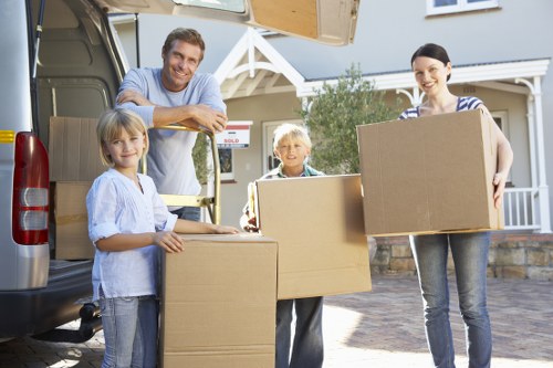 A family of four, including two children and two adults, are engaged in a house removal process outside a suburban property in Chiswick. They are unloading cardboard boxes of various sizes from the open rear of a large, dark grey removals van parked on a residential driveway. The boxes are plain, brown corrugated cardboard, and some are stacked on the ground while others are being carried by the family members. The children are smiling; the girl on the left holds a medium-sized box, while the young boy in the middle stands next to a larger box. The father, wearing a light blue shirt, is leaning on the open van door, and the mother, dressed in a striped top, is carrying a box towards the house. The background features a light-colored, two-storey house with white-framed windows, a small front garden with some greenery, and an overall bright, sunny atmosphere, consistent with typical house removal scenarios in the Chiswick area. This scene reflects professional house removals tailored to local residential areas, emphasizing careful handling and transportation of household belongings.