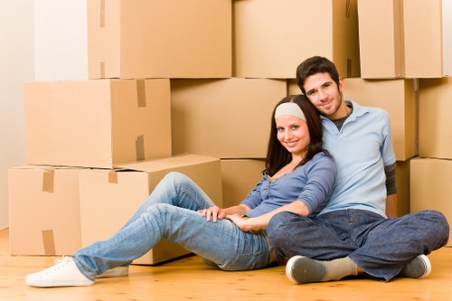 A young couple with light skin sitting on a wooden floor in a residential property, surrounded by multiple large cardboard moving boxes arranged in a semi-circle behind them. The woman, wearing a white headband, a blue long-sleeve top, and light blue jeans, is sitting with her back against the man's legs and smiling at the camera. The man, dressed in a light blue shirt and dark jeans, is sitting behind her with his legs crossed, leaning slightly toward her, and also smiling. The scene appears to be inside a house, possibly in a living room or hallway, with warm natural lighting illuminating the space. The boxes are sealed with packing tape, indicating a packing or moving process, consistent with house removal services offered in Chiswick. The overall setting suggests an active phase of relocating, with an emphasis on efficient packing and moving preparations by Chiswick Man with Van in the Chiswick area of West London, within the postcode W4.
