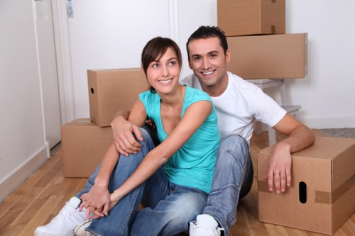 A smiling young couple with light skin, sitting on a light wooden floor inside a modern home, surrounded by packed cardboard moving boxes of various sizes. The woman has shoulder-length brown hair and is wearing a turquoise sleeveless top and blue jeans, while the man has short dark hair and is dressed in a white T-shirt and jeans. They are seated close together, with the woman leaning into the man's shoulder, both appearing relaxed and happy after moving. Behind them, there are more cardboard boxes stacked along a white wall, indicating a house relocation process. The environment suggests an interior area of a residential property, with natural light illuminating the scene, subtly supporting house removals and local moving services in Chiswick, London, near the postcode TW, connected with the website chiswickmanwithvan.co.uk and the page titled Complaints Procedure.