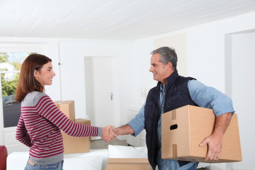 In a bright, modern residential interior with white walls and ceiling, a young woman wearing a red and white striped sweater and jeans is shaking hands with an older man dressed in a blue shirt and dark vest. The man is holding a medium-sized cardboard moving box in his left arm, while another similar box is positioned on a table or surface in front of him. The background features a visible door, a window allowing natural light into the room, and some additional cardboard boxes, indicating a house move process. The scene suggests a professional house removals or moving service, with the two individuals exchanging a gesture of agreement or greeting, against a clean and organized setting typical of a home in Chiswick or nearby areas in west London. The lighting is natural and evenly distributed, emphasizing the friendly interaction and the contents associated with a residential move.