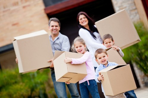 A family of four, including a man, woman, and two children, standing outdoors in front of a residential house, engaged in a moving activity. The man, positioned on the left, wears a light blue shirt and jeans, holding a large cardboard box. The woman, on the right, wears a white jacket and dark trousers, also holding a cardboard box. The young girl, with curly hair and dressed in a pink top, smiles while clutching a medium-sized cardboard box. Next to her, a boy in a blue shirt holds an open cardboard box, smiling towards the camera. All are standing on a paved driveway with a green lawn, and a partially visible brick house and garage are in the background, suggesting a house removal in Chiswick or nearby London. The scene is well-lit, with natural daylight, highlighting the cardboard boxes, their fabric and cardboard textures, and the casual attire suitable for a household move. The image conveys a family-oriented removals service environment, subtly associated with local household relocation activities.