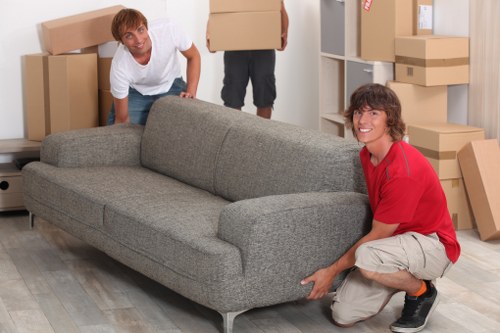 Two individuals are engaged in a household moving activity inside a room that appears to be part of a residential property, potentially in Chiswick, based on the context. The person on the left, wearing a white shirt and jeans, is crouching slightly behind a large, grey fabric sofa, which is positioned centrally in the room. The sofa has a smooth, textured upholstery, with wide armrests and a modern design. The person on the right, dressed in a red top and light-colored shorts, is kneeling beside the same sofa, holding its armrest to assist with lifting or moving it. The background features a storage area with white shelving units filled with uniformly packed cardboard boxes of various sizes, some with labels, and additional boxes stacked on shelves. The environment is well-lit, likely by natural or artificial light, emphasizing the clean, tidy space prepared for a house move. The scene reflects typical house removals involving careful handling of furniture and household items, consistent with services provided by Chiswick Man with Van in the local Chiswick area, with the scene set in a residential interior designed for efficient packing and relocation of personal belongings in the home. The scene visually underscores the careful, planned process of house moves or furniture relocation within the UK. 