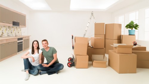 A young couple is seated on the light-colored wooden floor of a bright, modern interior room, surrounded by moving boxes made of brown cardboard, some stacked and others scattered. The woman has long dark hair, and is wearing a white top and blue jeans, while the man has short brown hair, wearing a green T-shirt and dark trousers; both are smiling at the camera. Behind them, large windows with white frames bring natural light into the space. To the right, a small green potted plant sits on a windowsill, adding a touch of greenery. A white foldable ladder leans against the wall near the boxes, indicating a recent or upcoming move. A black and red hand truck is partially visible behind the boxes. The room appears to be a kitchen or living area with modern cupboards, a beige backsplash, and a countertop on the left side. The scene suggests a home in Chiswick close to the W4 postcode, with the individuals involved in a house relocation process organized by Chiswick Man with Van, a professional removals service provider.