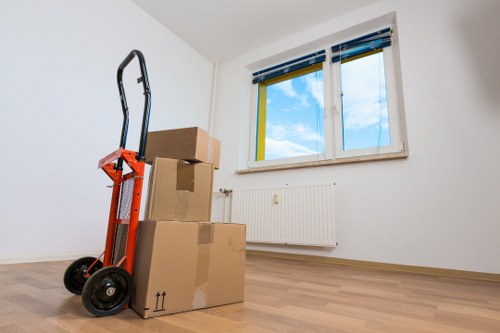 The image shows a minimally furnished, bright room with white walls and a light wooden floor, likely part of a residential property in Chiswick or nearby west London. In the foreground, there is a red hand truck with black wheels positioned upright, leaning against a pile of three cardboard moving boxes of varying sizes. The boxes are sealed with packing tape, and the top box is slightly smaller, stacked on larger ones with visible electrical symbols indicating it should be kept upright. Behind the boxes, a large double-glazed window with a white frame allows natural daylight to fill the space, revealing a clear blue sky with a few clouds outside. Below the window, a white radiator is mounted on the wall. The room appears tidy, with no other furniture or personal items visible, suggesting a moving or packing process, typical of house removals services offered in Chiswick or the surrounding areas.