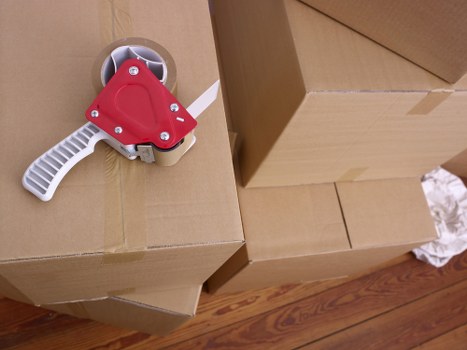 A close-up view of a cardboard box stacked on top of other boxes in a residential interior, with a red and white tape dispenser resting on the top box. The tape dispenser has a red plastic body, a white serrated cutting edge, and a white handle. The boxes are plain brown, unmarked, and appear to be used for packing household items during a house move, likely in Chiswick, west London. The background shows a wooden floor with a natural tone, typical of a UK home, and the environment appears well-lit, possibly by natural daylight. The image emphasizes the preparation and packing phase of a house removal service, illustrating the careful organization of personal belongings for a move within the residential property. The context and details reflect the typical process of packing household items using standard cardboard boxes and packing accessories during local removals in the Chiswick area.