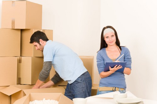 A young man and woman involved in house removals are seen in a room with white walls, stacking and unpacking cardboard boxes. The man, wearing a light blue t-shirt with grey sleeves and dark jeans, is bending over a box to pack or unpack items. The woman, dressed in a blue long-sleeved top and beige trousers, stands upright holding a clipboard and pen, looking towards the camera with a neutral expression. Behind them, several large, sealed cardboard boxes are stacked against the wall, indicating an ongoing move in a residential property, likely in or near Chiswick, as suggested by the website context. The scene is well-lit and minimalistic, with a white table or surface in the foreground holding additional small items and packing materials, emphasizing a professional but domestic setting typical for house removal services in London or nearby areas.