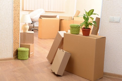 The image displays a room in a residential property, likely in Chiswick, with several moving boxes and household items in the process of being packed or moved. In the foreground, a large, rectangular cardboard box is positioned on the wooden floor, with a smaller box leaning against it. A green, woven storage basket is placed on top of the large box, alongside a potted houseplant with broad, green leaves situated on top of a medium-sized cardboard box. To the left, a smaller, round, green fabric storage container is on the floor near a light wooden side table. In the background, additional cardboard boxes are stacked, some closed and others slightly open, revealing packing materials. The room features a cozy, well-lit atmosphere with soft natural light, and visible interior details include a textured wallpapered wall, a pale side lamp, and a partially visible sleeping area with a bed and a bedside lamp. The scene suggests a house removal or relocation process, consistent with services offered by Chiswick Man with Van in the Chiswick area.