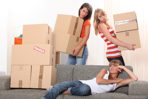 Two young women are engaged in a house moving activity inside a residential living room. One woman, wearing an orange top and jeans, is standing behind a stack of large cardboard boxes, lifting and carrying one of the boxes, which appears to contain household items. The other woman, dressed in a red and white striped top and dark shorts, is holding a smaller cardboard box and is positioned beside the sofa, preparing to move it. In front of the sofa, a young boy with blonde hair, wearing a white t-shirt and blue jeans, is lying on his front with his head resting on his hand and looking towards the camera. The background shows a light wall and a doorway, with natural lighting illuminating the scene. The boxes vary in size, some labeled with 'Fragile,' indicating delicate contents, and are arranged in a semi-organized manner throughout the room, which appears prepared for a domestic move. This scene reflects typical house removal activities, with attention to safe handling and organization, and subtly references local residential settings consistent with Chiswick or nearby areas close to W4 postal code.