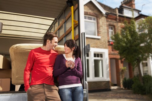 Two individuals, a man in a red long-sleeve shirt and a woman in a purple sweater, are standing outside the back of a removals vehicle on a residential street in Chiswick, London, near the postcode area W4. They are smiling and engaged in conversation, with the man leaning slightly towards the woman, who is holding a cup of coffee or tea. The vehicle's open rear reveals packed cardboard boxes and furniture, including a beige fabric sofa resting on the vehicle's floor. The background shows a row of brick terraced houses with bay windows, white window frames, and small front gardens with trees and shrubs, typical of a suburban London area. Natural daylight illuminates the scene, emphasizing the collaborative and positive atmosphere of a house relocation. The image reflects home moving activities carried out by Chiswick Man with Van, highlighting their residential removals service for local communities in W4.