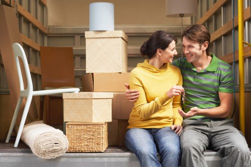 A man and woman sit on the edge of a moving box-filled storage area, surrounded by various household items such as cardboard boxes, wicker baskets, and a rolled-up beige rug, suggesting preparations for a house move within Chiswick. The woman, wearing a yellow hoodie and blue jeans, has dark hair tied back and is smiling while holding a small object, possibly key or remote, in her hand. The man, wearing a green and navy striped polo shirt and grey trousers, has light brown hair and is smiling back at her. Behind them are stacked cardboard boxes, some with textured surface finishes, and a large beige storage box on top of the stack, with a white lamp shade resting on the highest box. To the left, a white plastic chair and a second rolled-up rug are visible. The scene is indoors, likely in a home or storage unit, with wooden shelving to the right and a neutral-toned background, indicating a residential setting typical of a house removal service in the Chiswick area, as managed by Chiswick Man with Van.