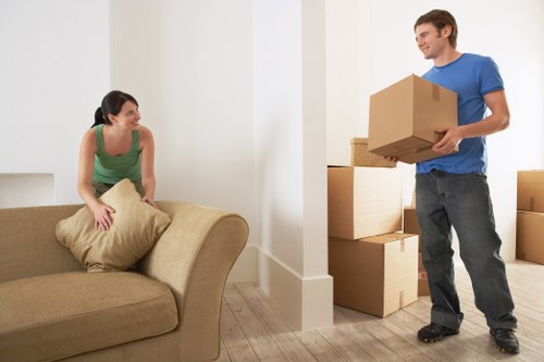In a well-lit room with light-colored walls and a wooden floor, a woman wearing a green top is seated on a beige upholstered sofa, holding a large beige cushion and smiling at a man. The man, dressed in a blue t-shirt and dark jeans, is standing beside a doorway, holding a medium-sized cardboard box. Several other cardboard boxes of various sizes are stacked nearby, some on the wooden floor and others on a small shelf or ledge. The scene captures an active moment of house removals, with the man appearing to be in the process of moving items into or out of the room, suggesting a residential setting typical of Chiswick. The environment suggests a professional removals service, such as Chiswick Man with Van, operating within or near the London suburb, with a focus on careful handling of household belongings during relocation.