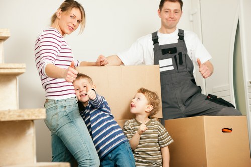 A family of four in a well-lit interior space, preparing for a house move. The mother, wearing a striped long-sleeve top and jeans, is standing to the left, giving a thumbs-up gesture. Two children, a boy in a blue and white striped shirt and a girl in a beige and black striped top, are positioned in front of her, smiling and looking at the camera, with the boy giving a thumbs-up. Behind them, a man dressed in grey work overalls and a white T-shirt is standing near a stack of cardboard moving boxes, giving a thumbs-up. The background includes a staircase on the left and a white wall with a doorframe on the right, suggesting a residential setting. The scene depicts a cheerful moment of family readiness for a home relocation service, with the boxes indicating packing activity typical of house removals in the Chiswick area, subtly supporting local SEO for Chiswick Man with Van.