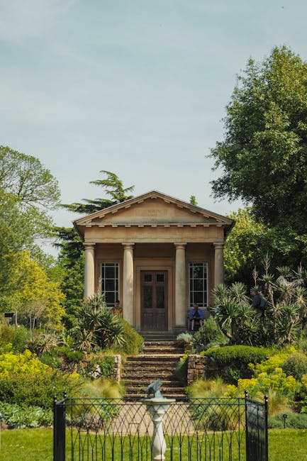 The image depicts the front view of a classical-style building surrounded by lush greenery, including tall trees and well-maintained shrubs. The building features a symmetrical facade with four column
