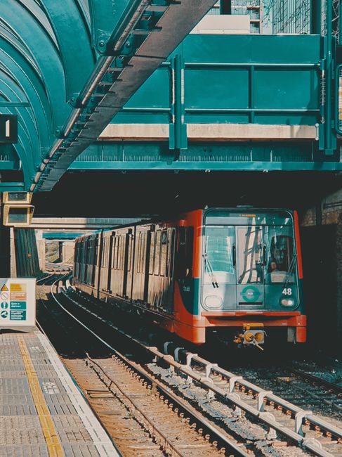 A modern commuter train with a sleek, blue exterior traveling along railway tracks during daylight hours, with overhead electrical wires and supporting steel structures visible above the train. The tr