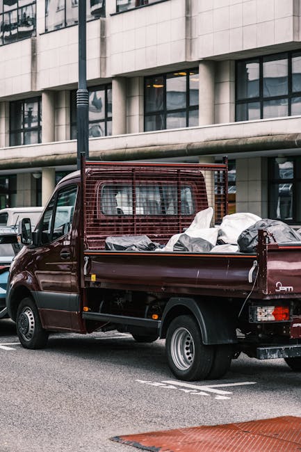 A dark red mini truck parked on a city street in front of a modern office building with large glass windows and concrete panels. The truck's flatbed is loaded with various household items, including c